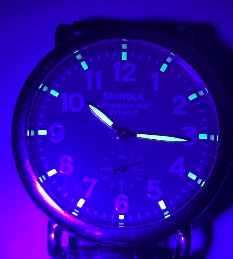 Może przedstawiać: Zbliżenie tarczy zegarka Shinola Argonite-1069 podświetlonej niebieskim światłem. Zegarek ma świecące wskaz&oacute;wki i znaczniki, z widocznym tekstem "SHINOLA Argonite-1069 DETROIT". Tarcza zegarka wyświetla liczby od 1 do 12.