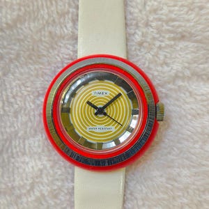 May include: A round Timex wristwatch with a bright red bezel and a yellow dial featuring concentric circles. The watch has black hands, a silver-toned metallic inner ring, and a smooth white leather strap. The dial text reads Timex and Water Resistant.