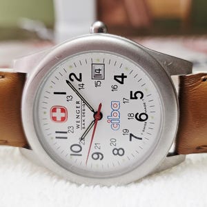 May include: A silver-toned Wenger watch with a white face, black numerals, and a red second hand. The watch features a brown leather strap and a date display at the 3 o'clock position. The watch face includes the text "WENGER S.A.K. DESIGN" and "ciba".