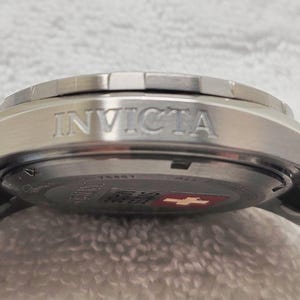 Puede incluir: Primer plano de un reloj Invicta plateado. El reloj presenta una correa de acero inoxidable y una caja plateada con el nombre de la marca "INVICTA" grabado en relieve en el lateral. La esfera del reloj es visible, con un emblema rojo y blanco.