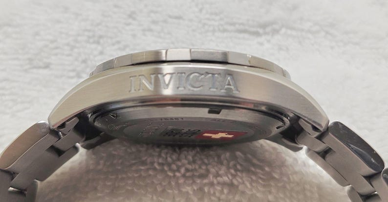 Puede incluir: Primer plano de un reloj Invicta plateado. El reloj presenta una correa de acero inoxidable y una caja plateada con el nombre de la marca "INVICTA" grabado en relieve en el lateral. La esfera del reloj es visible, con un emblema rojo y blanco.