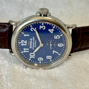 Może przedstawiać: Zbliżenie zegarka Shinola Argonite-1069 Detroit z niebieską tarczą i białymi cyframi. Zegarek ma srebrną kopertę i brązowy sk&oacute;rzany pasek. Tarcza zegarka zawiera małą tarczę sekundową.