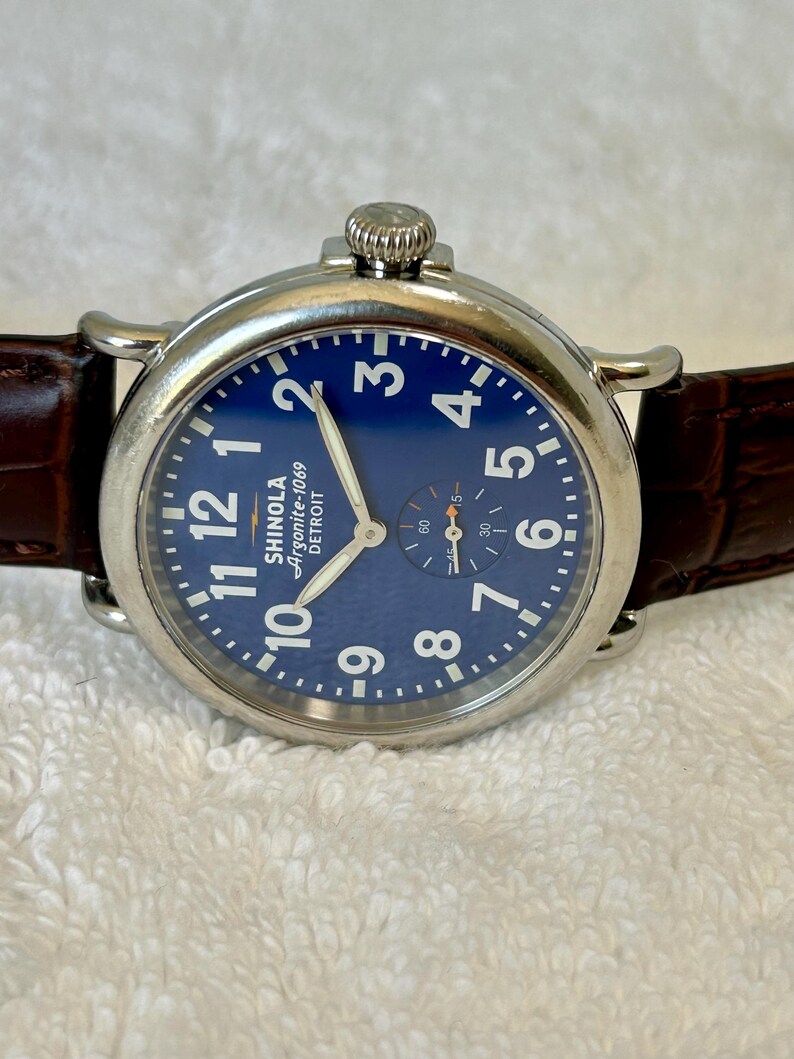 Może przedstawiać: Zbliżenie zegarka Shinola Argonite-1069 Detroit z niebieską tarczą i białymi cyframi. Zegarek ma srebrną kopertę i brązowy sk&oacute;rzany pasek. Tarcza zegarka zawiera małą tarczę sekundową.