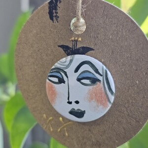 May include: A circular brown tag featuring a hand-painted face. The face has blue eyes, peach cheeks, and black details on a white background. A gold string is attached. The background shows blurred green leaves.