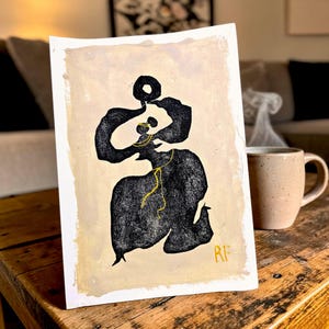 Puede incluir: Impresión artística abstracta que presenta una figura negra con detalles dorados sobre un fondo beige. La obra de arte está firmada con las iniciales "RF" en dorado. Una taza de cerámica con vapor saliendo de ella se encuentra cerca sobre una mesa de madera.