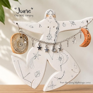 Könnte beinhalten: Ein weißer Wandbehang aus Ton mit dem Titel June aus der Serie Living Mythologies. Die Figur zeigt schwarze Strichzeichnungen von Himmelsymbolen, eine silberne Kette mit Sternanhängern, ein kreisförmiges Holzstück und eine Mondsichel.