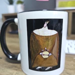 Könnte beinhalten: Weiße Keramiktasse mit schwarzem Henkel und Rand. Die Tasse zeigt eine farbenfrohe Illustration einer Figur mit Krone und goldfarbenem Gewand, die eine juwelenbesetzte Kugel hält. Der Hintergrund ist schwarz.