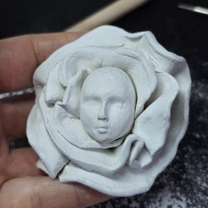 Könnte beinhalten: Eine weiße Ton-Skulptur einer Rose mit einem menschlichen Gesicht in der Mitte. Die Rosenblätter sind detailliert und umgeben das Gesicht, das zarte Züge aufweist. Die Skulptur ist handgefertigt und scheint ein Werk in Arbeit zu sein.