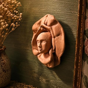 May include: A terracotta wall sculpture depicting a serene female face emerging from draped fabric. The sculpture is mounted on a dark green wall, with a vase of dried flowers and a framed picture in the background.