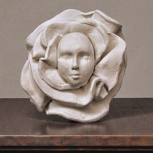 Puede incluir: Una escultura blanca de un rostro emergiendo de una rosa. El rostro tiene los ojos cerrados y rasgos sutiles. La escultura está sobre una superficie de madera oscura, con un fondo de color neutro.