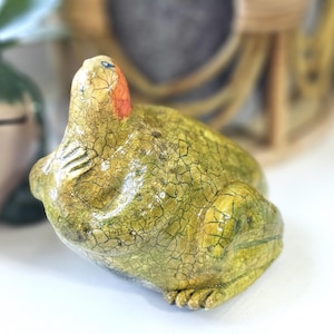 Könnte beinhalten: Keramikfroschfigur mit strukturierter, rissiger Oberfläche in Grün- und Gelbtönen. Der Frosch hat einen roten und blauen Akzent auf dem Kopf. Die Figur ist ein dekorativer Artikel.