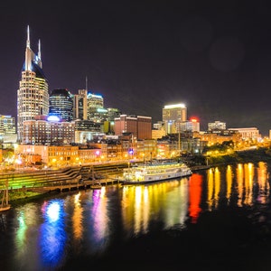 Może przedstawiać: Nocny widok na panoramę Nashville z ikonicznym Ryman Auditorium w tle. Miasto jest oświetlone światłami, kt&oacute;re odbijają się w wodzie. Ł&oacute;dź jest zacumowana na pierwszym planie.