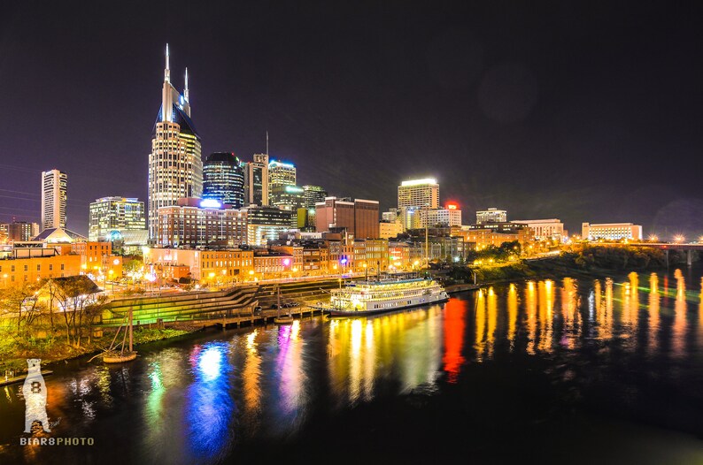 Może przedstawiać: Nocny widok na panoramę Nashville z ikonicznym Ryman Auditorium w tle. Miasto jest oświetlone światłami, kt&oacute;re odbijają się w wodzie. Ł&oacute;dź jest zacumowana na pierwszym planie.