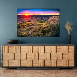 May include: A colorful sunset over a canyon landscape with a blue sky and pink, orange, and yellow clouds. The canyon is covered in green grass and trees. The image is framed and hanging on a blue wall above a wooden cabinet.