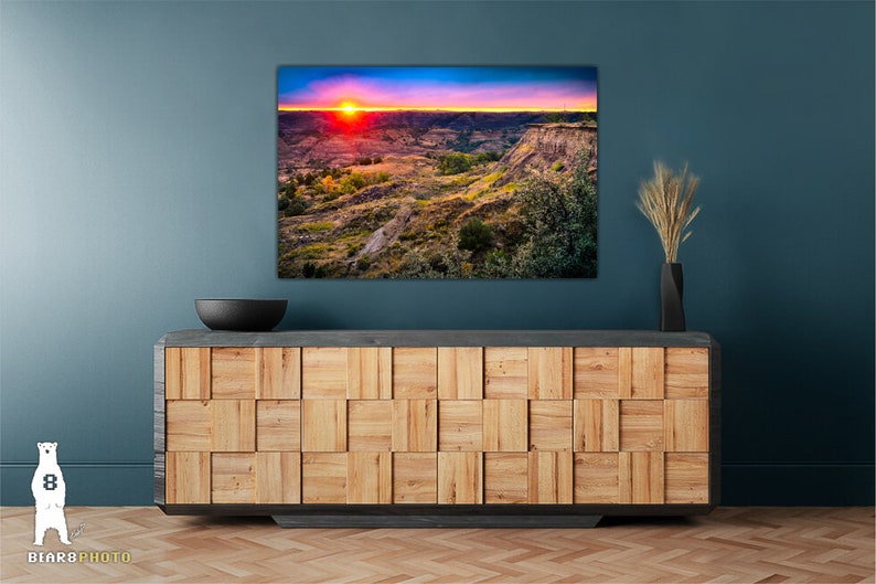 May include: A colorful sunset over a canyon landscape with a blue sky and pink, orange, and yellow clouds. The canyon is covered in green grass and trees. The image is framed and hanging on a blue wall above a wooden cabinet.