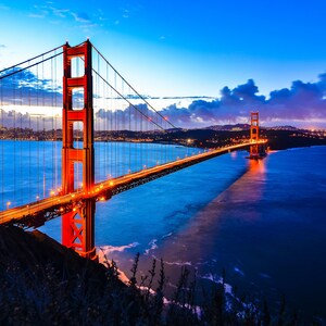 May include: A view of the Golden Gate Bridge in San Francisco, California at dusk. The bridge is lit up with orange lights and the water is a deep blue. The city skyline is visible in the distance.