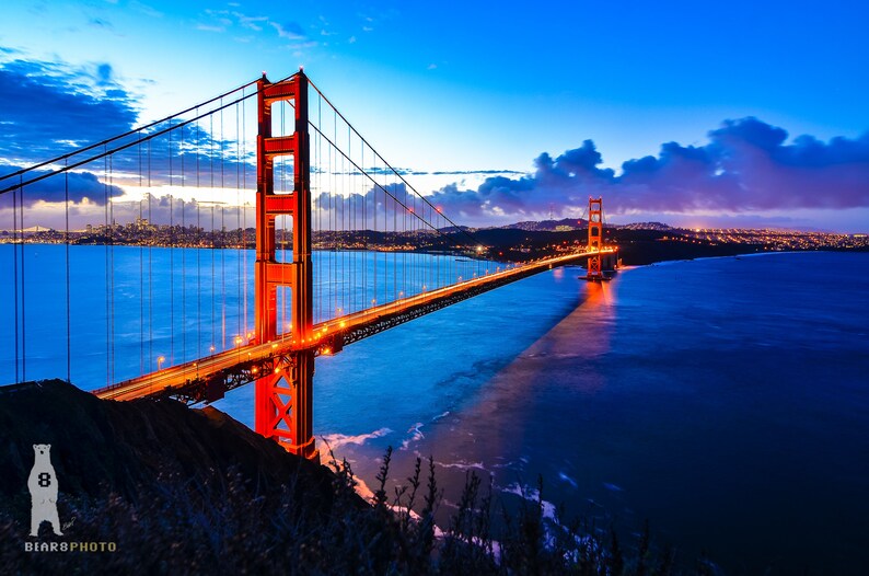 May include: A view of the Golden Gate Bridge in San Francisco, California at dusk. The bridge is lit up with orange lights and the water is a deep blue. The city skyline is visible in the distance.