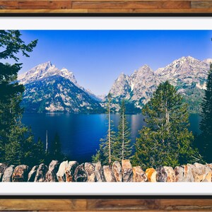 May include: A scenic view of a mountain range with snow-capped peaks reflected in a still lake. The foreground features a stone wall and lush green trees.