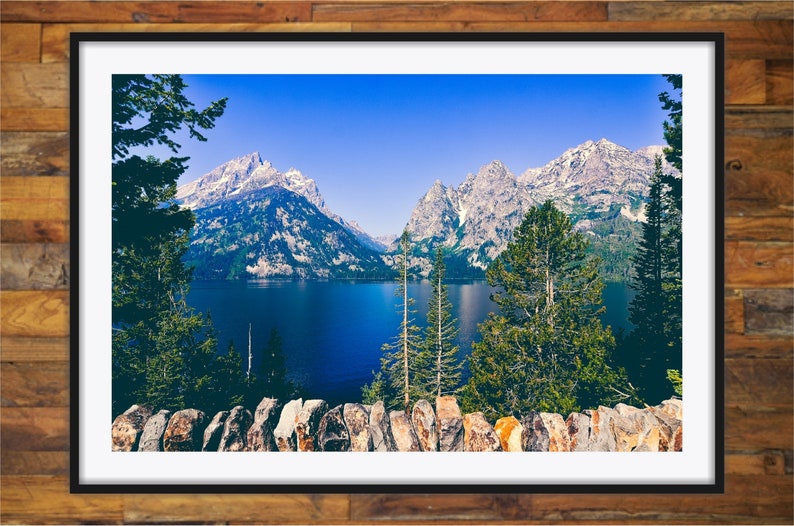 May include: A scenic view of a mountain range with snow-capped peaks reflected in a still lake. The foreground features a stone wall and lush green trees.