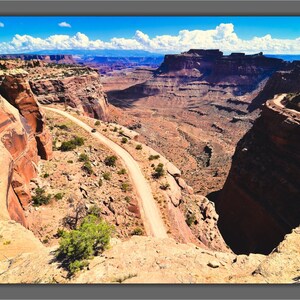 May include: A winding dirt road snakes through a canyon in a desert landscape. The road is surrounded by red rock cliffs and the sky is blue with white clouds.