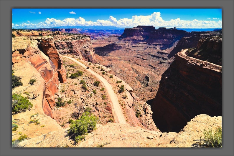 May include: A winding dirt road snakes through a canyon in a desert landscape. The road is surrounded by red rock cliffs and the sky is blue with white clouds.