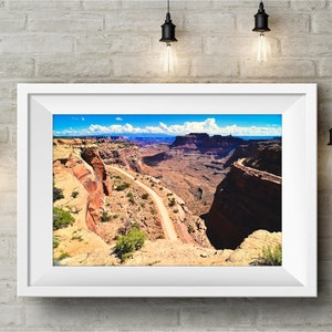 May include: A scenic photograph of a winding dirt road through a canyon in a desert landscape. The road leads to a distant mountain range under a bright blue sky with white clouds.