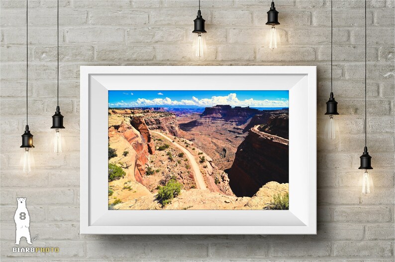 May include: A scenic photograph of a winding dirt road through a canyon in a desert landscape. The road leads to a distant mountain range under a bright blue sky with white clouds.