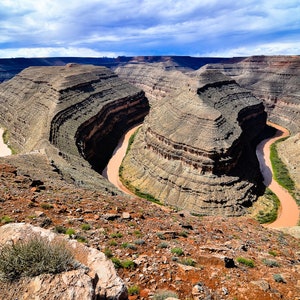 May include: An aerial view of a winding river carving through a canyon in a desert landscape. The river is a light brown color and the canyon walls are a mix of brown, tan, and gray. The sky is a bright blue with white clouds.