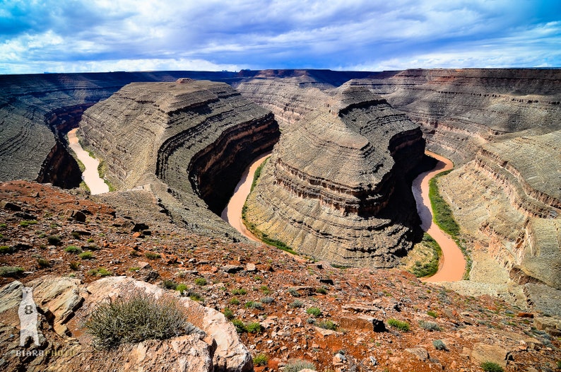 May include: An aerial view of a winding river carving through a canyon in a desert landscape. The river is a light brown color and the canyon walls are a mix of brown, tan, and gray. The sky is a bright blue with white clouds.