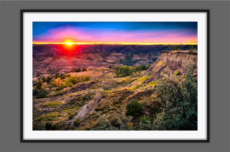 May include: A vibrant sunset over a rugged landscape with a deep canyon and rolling hills. The sky is a mix of pink, orange, and blue, with the sun setting in the distance.