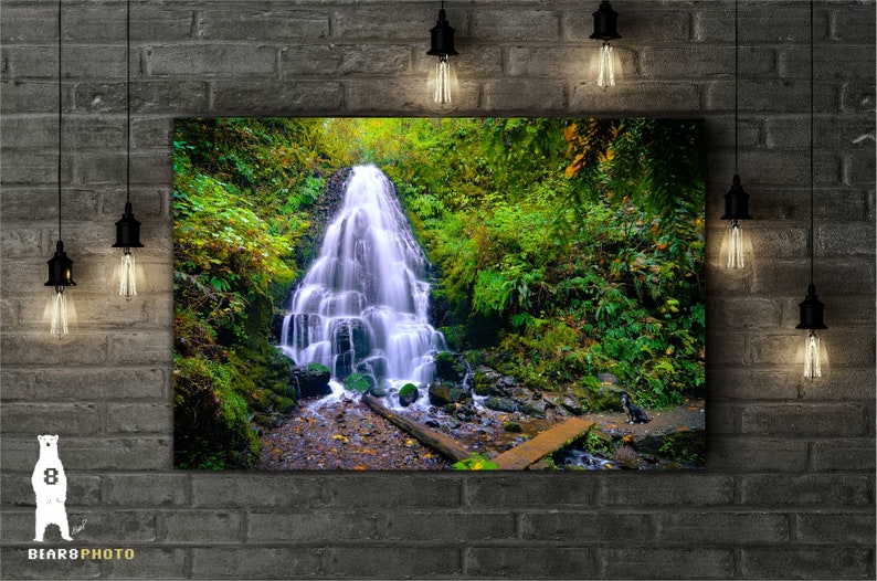 May include: A lush green forest scene with a cascading waterfall flowing over moss-covered rocks. The water is clear and blue, and the surrounding foliage is vibrant green.