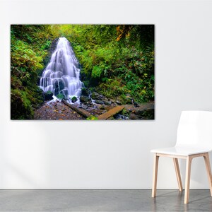 May include: A cascading waterfall flows through a lush green forest. The water is white and the surrounding foliage is a vibrant green. A wooden bridge crosses the stream below the waterfall.
