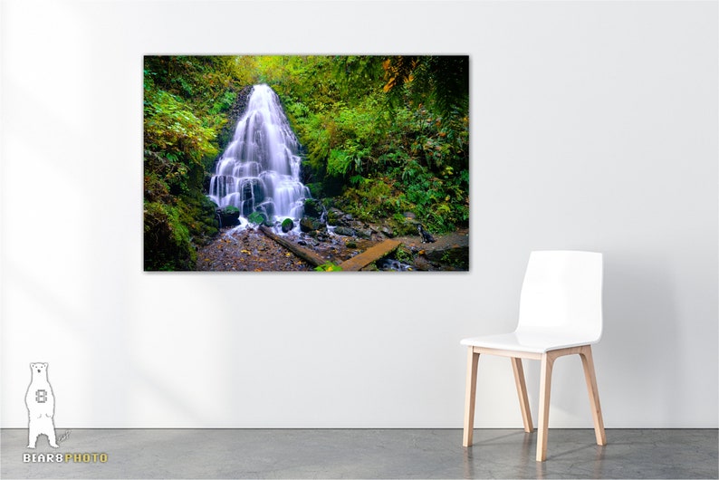 May include: A cascading waterfall flows through a lush green forest. The water is white and the surrounding foliage is a vibrant green. A wooden bridge crosses the stream below the waterfall.