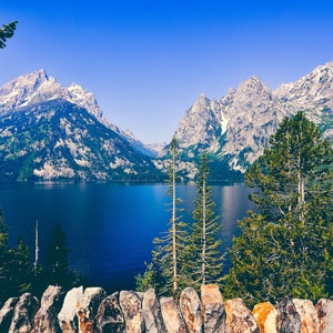 May include: A scenic view of a lake surrounded by snow-capped mountains and lush green trees. The water is a deep blue and the sky is a clear blue.