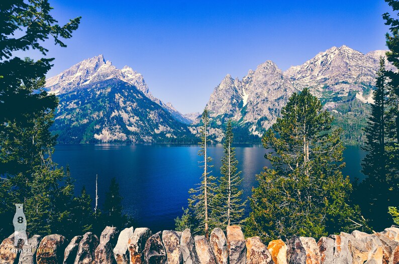 May include: A scenic view of a lake surrounded by snow-capped mountains and lush green trees. The water is a deep blue and the sky is a clear blue.