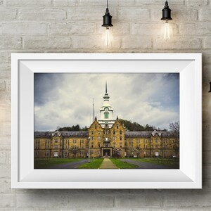 May include: A framed photograph of a large, yellow brick building with a green roof and a tall tower. The building has many windows and a long, symmetrical facade. The sky is cloudy and gray.