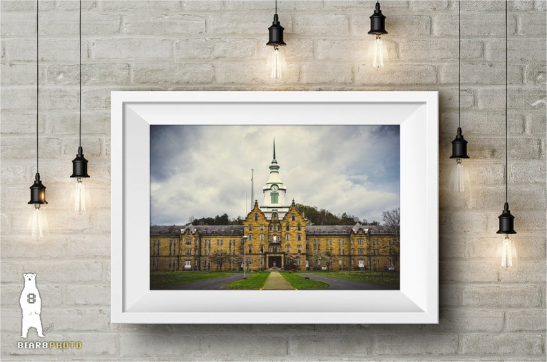 May include: A framed photograph of a large, yellow brick building with a green roof and a tall tower. The building has many windows and a long, symmetrical facade. The sky is cloudy and gray.