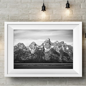 May include: A black and white photograph of a mountain range with snow-capped peaks. The mountains are reflected in a still body of water in the foreground.