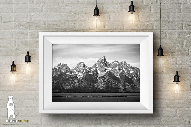 May include: A black and white photograph of a mountain range with snow-capped peaks. The mountains are reflected in a still body of water in the foreground.