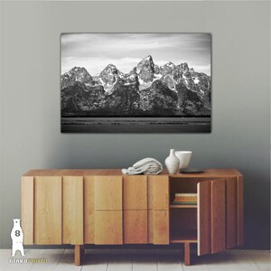 May include: Black and white photograph of a mountain range with snow-capped peaks. The mountains are reflected in a still body of water in the foreground. The image is framed and hanging on a wall above a wooden cabinet.