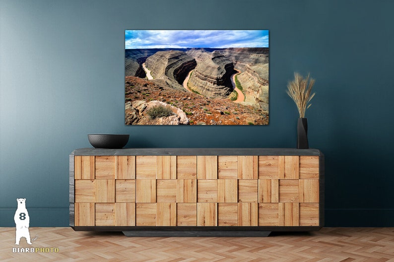 May include: A photograph of a scenic landscape featuring a winding river flowing through a canyon. The canyon walls are a mix of brown, red, and gray rock formations. The sky is blue with white clouds.