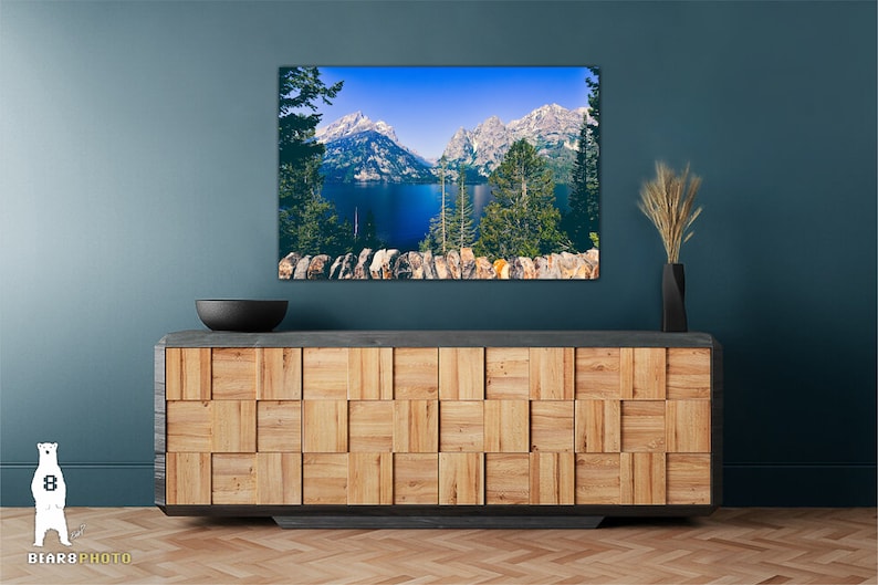 May include: A framed photograph of a mountain lake with snow-capped peaks in the background. The lake is a deep blue color and the mountains are a light blue and white. The photo is hanging on a teal wall above a wooden cabinet.