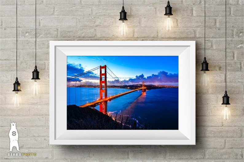 May include: A framed photograph of the Golden Gate Bridge in San Francisco, California. The bridge is lit up at night, with the city lights visible in the distance. The sky is a deep blue with pink and purple clouds.