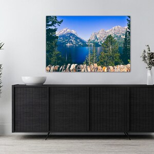 May include: A framed photograph of a mountain lake with snow-capped peaks in the background. The lake is a deep blue color and the mountains are a light blue and white. The photo is framed in a black frame and is hanging on a white wall.