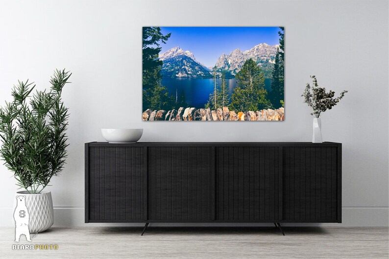 May include: A framed photograph of a mountain lake with snow-capped peaks in the background. The lake is a deep blue color and the mountains are a light blue and white. The photo is framed in a black frame and is hanging on a white wall.