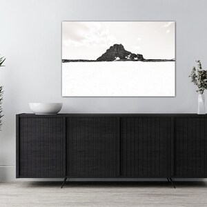 May include: A sepia-toned landscape photograph featuring a mountain against a cloudy sky. The artwork is displayed above a black cabinet with a white bowl and a vase of flowers. A potted plant is on the left.