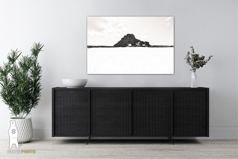 May include: A sepia-toned landscape photograph featuring a mountain against a cloudy sky. The artwork is displayed above a black cabinet with a white bowl and a vase of flowers. A potted plant is on the left.