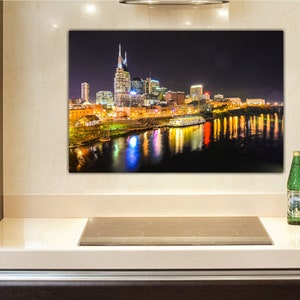Może przedstawiać: Nocne zdjęcie panoramy Nashville w stanie Tennessee, z rzeką Cumberland w pierwszym planie. Sylwetka miasta jest oświetlona światłami, a rzeka odbija światła. Ł&oacute;dź jest zacumowana w rzece.