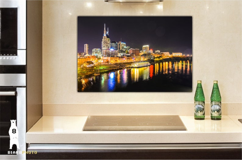 Może przedstawiać: Nocne zdjęcie panoramy Nashville w stanie Tennessee, z rzeką Cumberland w pierwszym planie. Sylwetka miasta jest oświetlona światłami, a rzeka odbija światła. Ł&oacute;dź jest zacumowana w rzece.