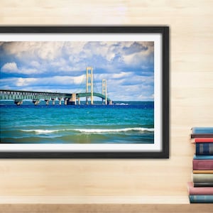 May include: A framed photograph of the Mackinac Bridge in Michigan. The bridge is a suspension bridge with a green roadway and yellow towers. The bridge is over a body of water with blue water and white waves. The sky is blue with white clouds.